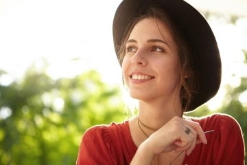Smiling woman wearing a hat outdoors, looking to the side in natural sunlight with a relaxed and happy expression.