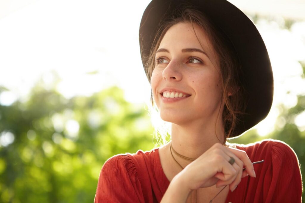 Smiling woman wearing a hat outdoors, looking to the side in natural sunlight with a relaxed and happy expression.