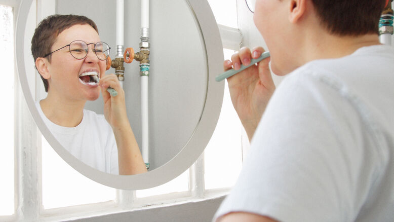 Person brushing their teeth with a toothbrush while looking into a bathroom mirror.